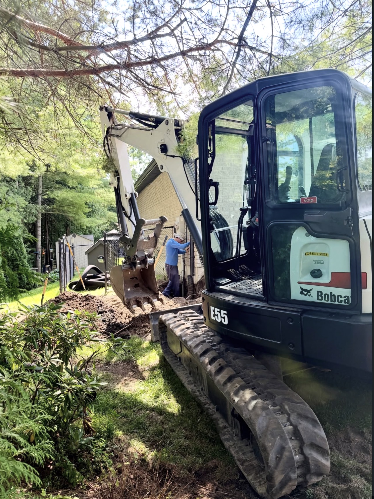 Bobcat E55 mini excavator digging a trench beside a brick house while a worker checks the line